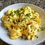 A close-up of creamy, fluffy scrambled egg served on a white plate and garnished generously with chopped fresh chives.