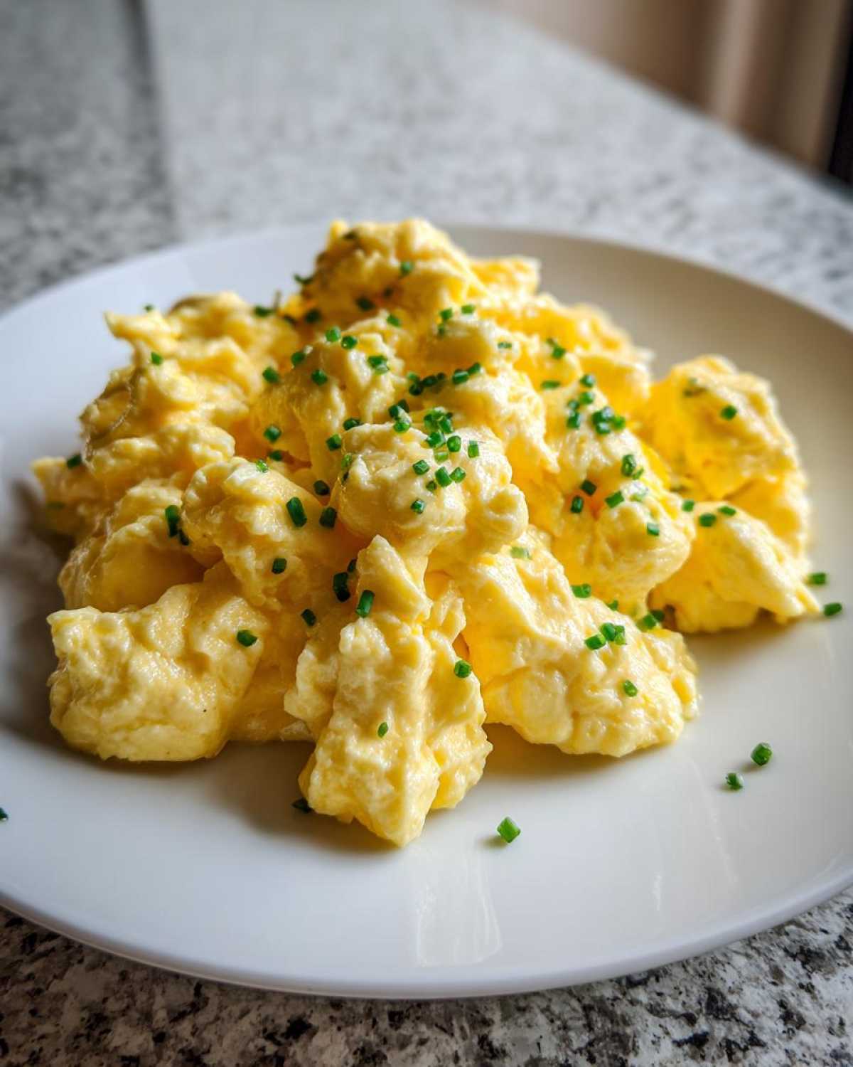 A close-up of creamy, soft scrambled egg served on a white plate and topped with finely chopped green chives.