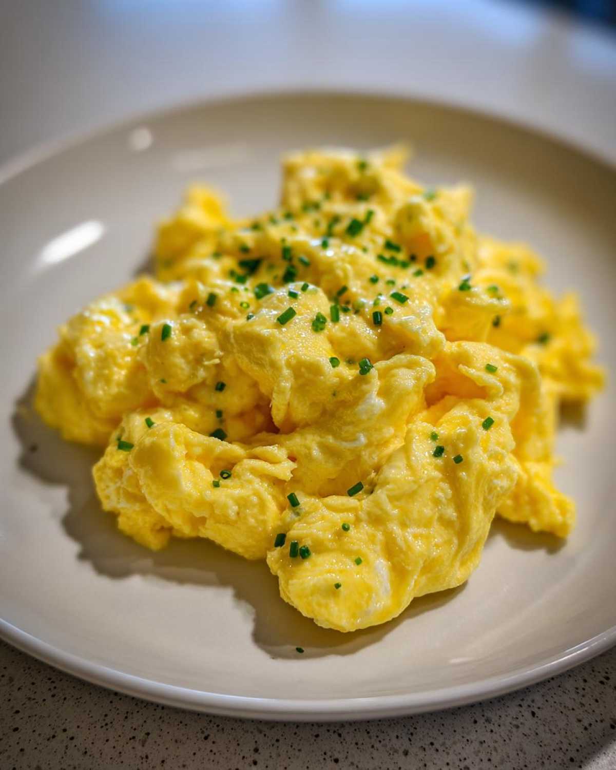 A close-up of creamy, fluffy scrambled egg piled high on a light plate, garnished with fresh chopped chives.