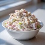 A close-up of a white bowl filled with creamy ham salad recipe, featuring diced pink ham and green celery, topped with cracked black pepper.