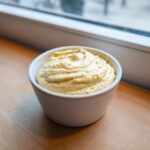 A small white bowl filled with thick, creamy Low Carb Garlic Aioli, set on a wooden surface near a window.