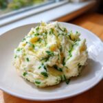 A serving of creamy Mashed Potatoes topped with melted brown butter and fresh green chives on a white plate.