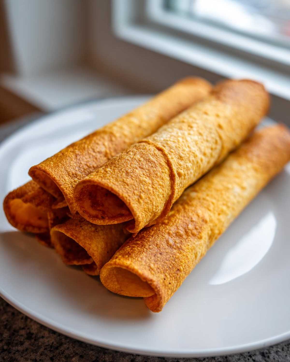 A stack of golden brown, crispy Chicken Taquitos resting on a white plate near a window.