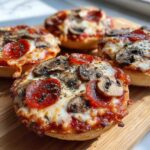 Close-up of a crispy English muffin pizza topped with melted cheese, pepperoni, mushrooms, and herbs.