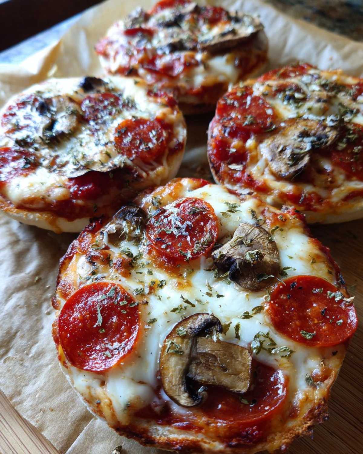 Close-up of four freshly baked English muffin pizzas topped with melted cheese, pepperoni, mushrooms, and herbs.