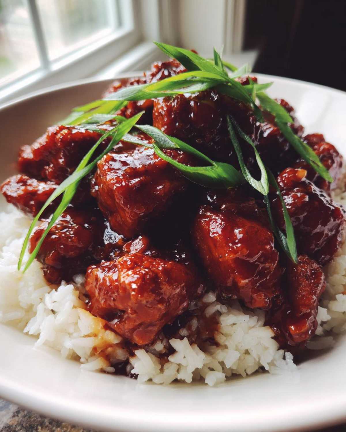 Close-up of saucy Crockpot Bourbon Chicken pieces served over fluffy white rice and garnished with sliced green onions.