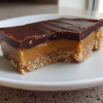 A close-up of a layered Date Snickers Bar slice featuring a crumbly base, thick caramel, and dark chocolate topping.