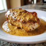 A juicy, pan-seared chicken breast smothered in a creamy, speckled dijon chicken sauce on a white plate.