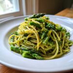 A close-up of a white bowl filled with Earth Day Dinner Pasta, featuring spaghetti tossed in green pesto and topped with bright green asparagus pieces.