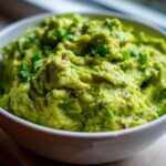 Close-up of a white bowl filled with chunky, bright green Easy Guacamole Recipe From Scratch, garnished with cilantro.