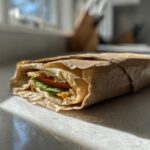 A single Eco Friendly Picnic Sandwich Wrap made with a tortilla, lettuce, tomato, and cheese, partially wrapped in brown paper.