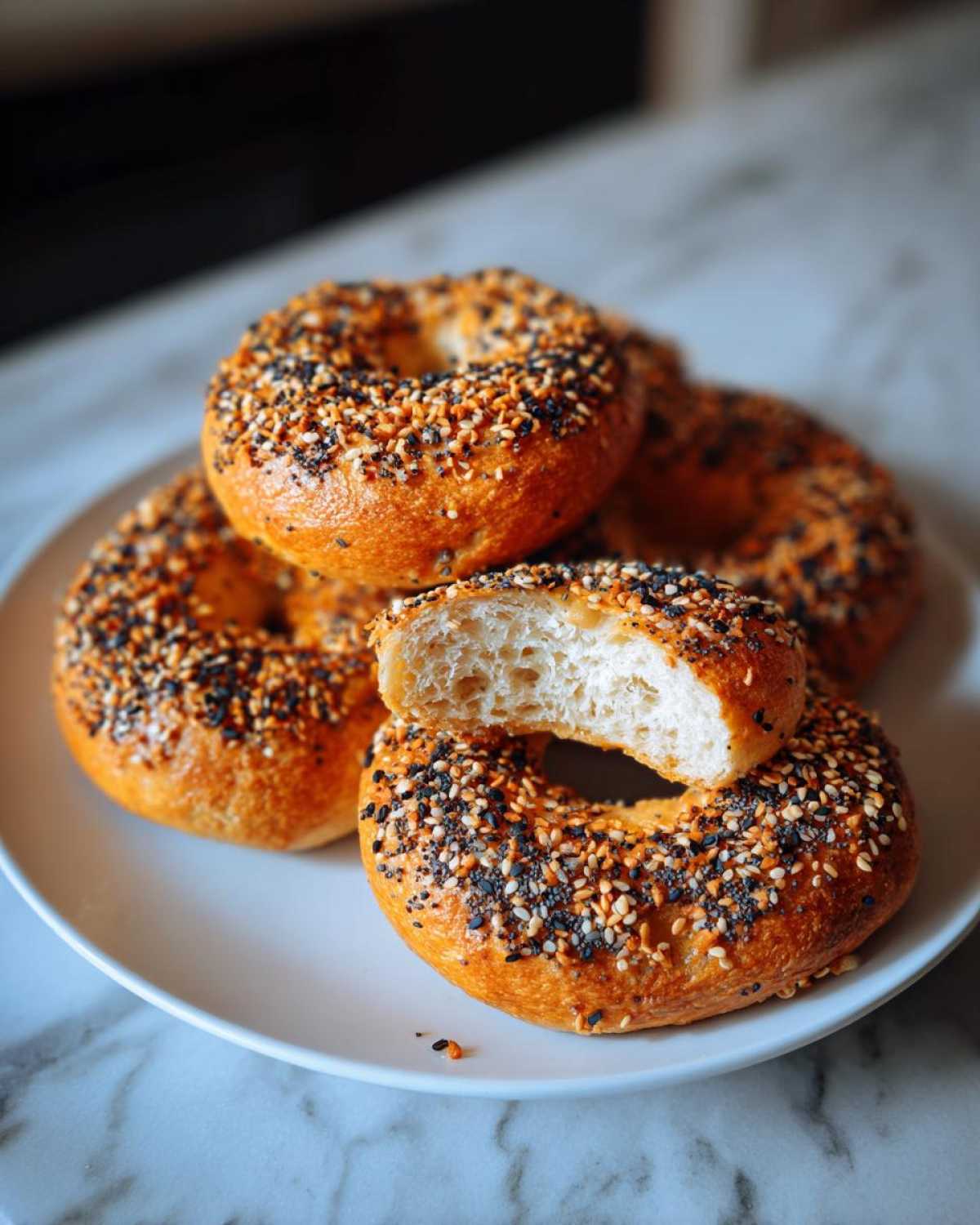 A stack of golden brown sourdough discard bagels topped with everything seasoning, one bagel is sliced open showing the airy interior.