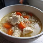 A close-up of a white bowl filled with homemade chicken and rice soup, featuring chunks of white chicken meat and bright orange carrot slices.
