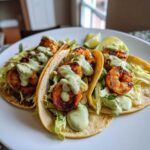 Three delicious Shrimp Tacos filled with seasoned shrimp, shredded lettuce, and drizzled with a light green creamy sauce.