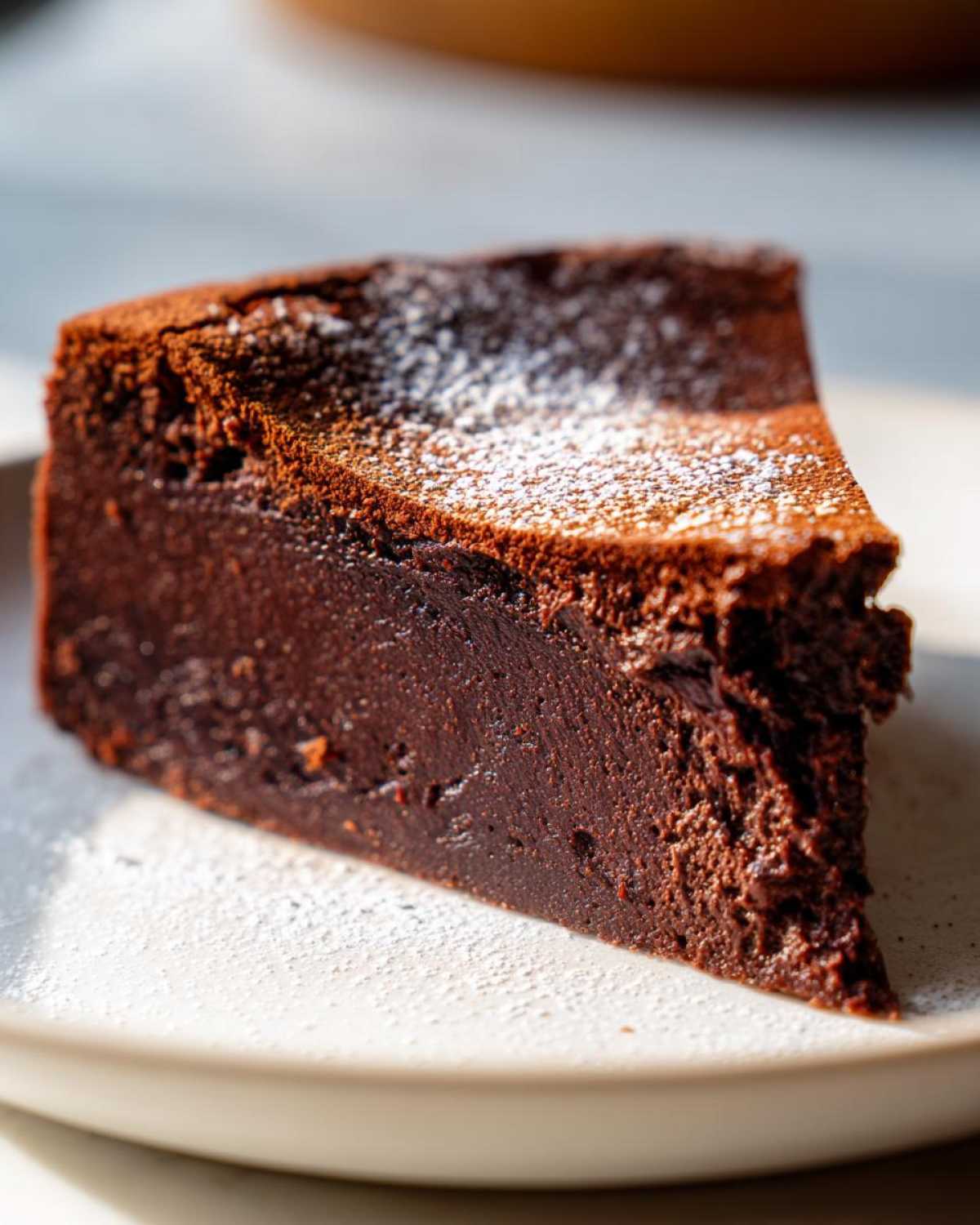 A close-up of a dense, dark slice of flourless chocolate cake dusted lightly with powdered sugar on a light plate.