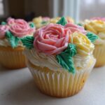 Close-up of a vanilla cupcake topped with buttercream frosting roses in pink and yellow, decorated with green leaves. Perfect Flower Cupcakes for Mothers Day.