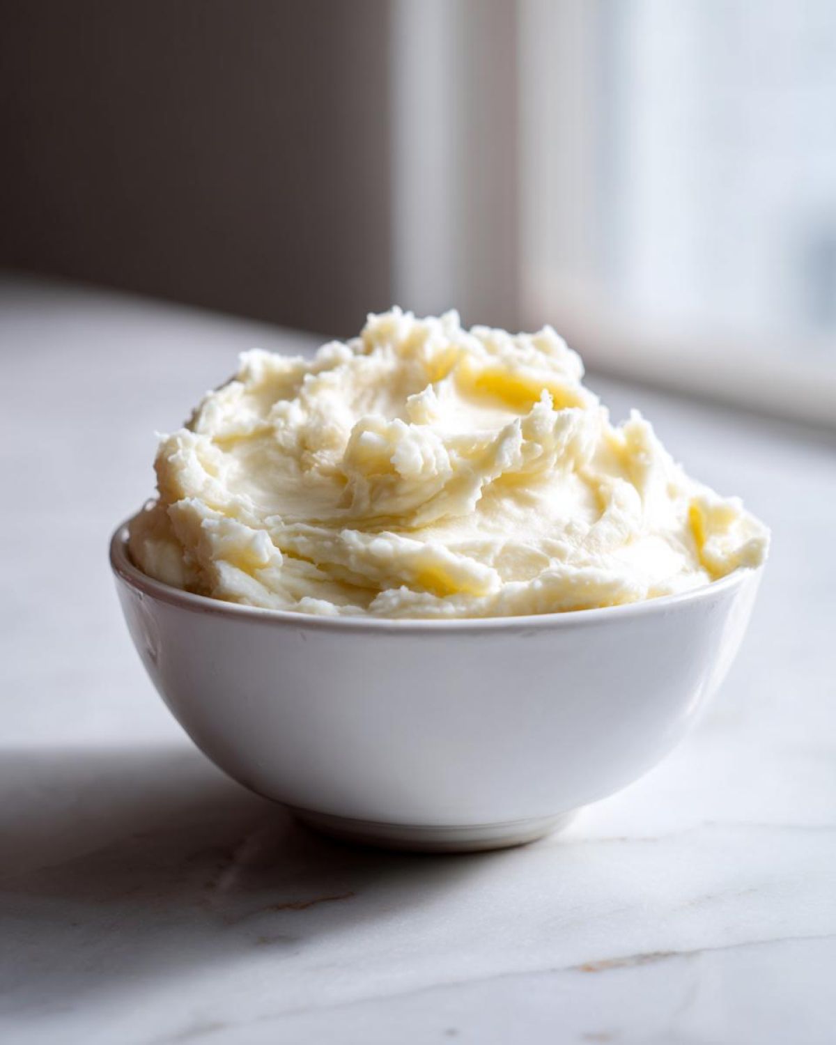 A close-up of creamy, fluffy mashed potatoes served in a white bowl on a marble surface.