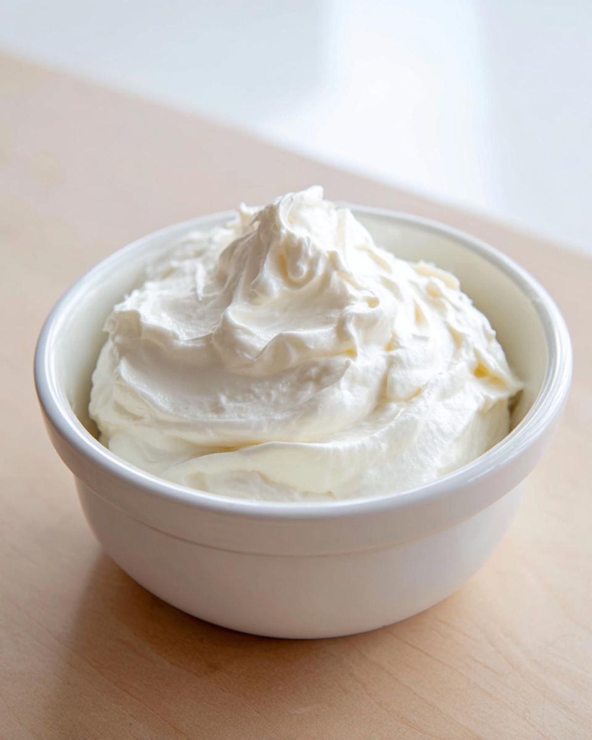 A close-up of fluffy white fruit dip swirled high in a small white bowl on a light wooden surface.