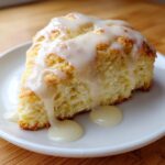 A close-up of a fluffy lemon scone topped with thick, dripping tangy glaze, served on a white plate.