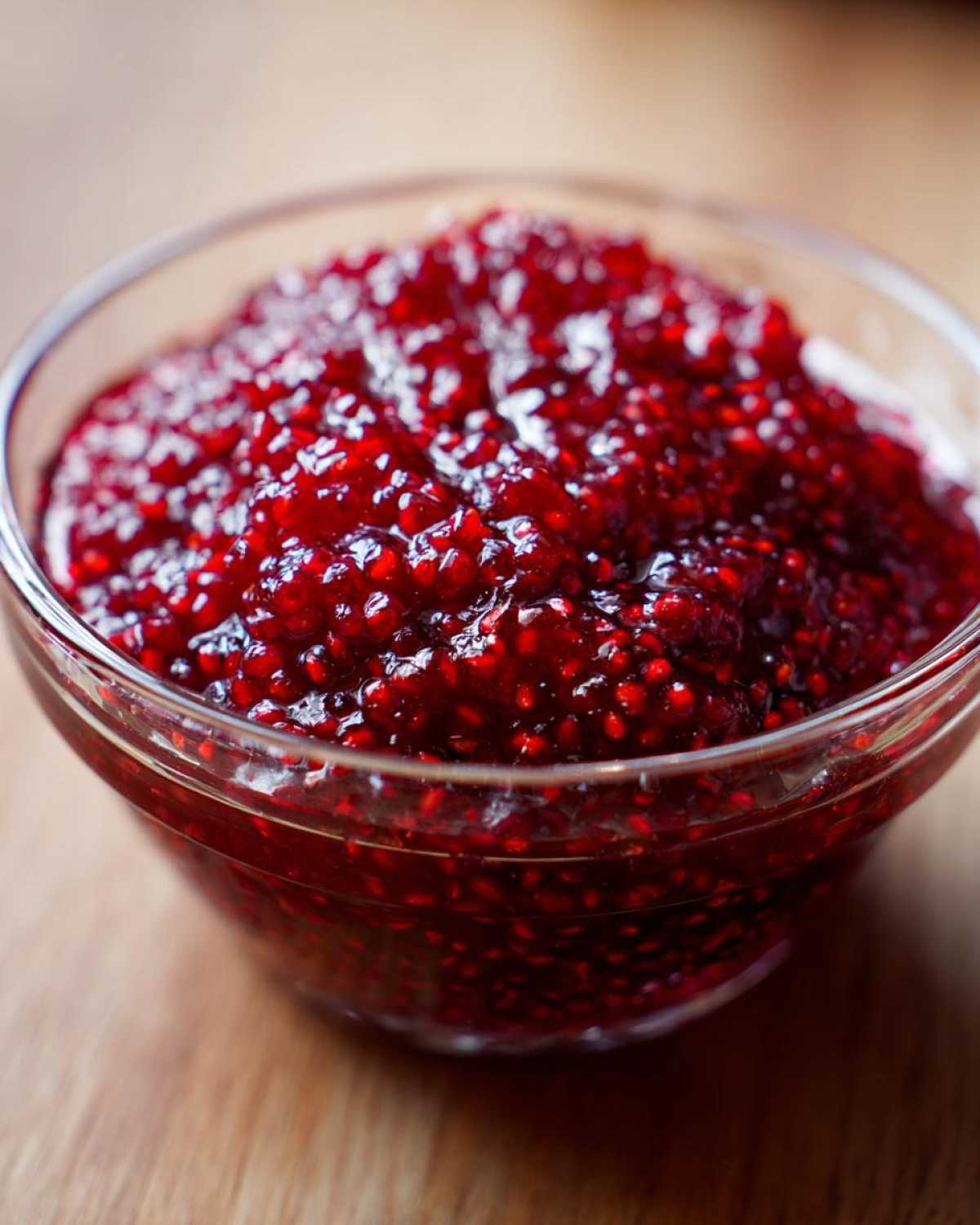 A close-up, vibrant image of homemade raspberry jam, showing the texture of seeds and fruit, served in a clear glass bowl.