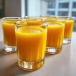Six small glasses filled with bright orange Ginger Lemon Turmeric Shots lined up on a light surface.