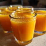Close-up of several small glasses filled with bright orange Ginger Lemon Turmeric Shots lined up on a counter.