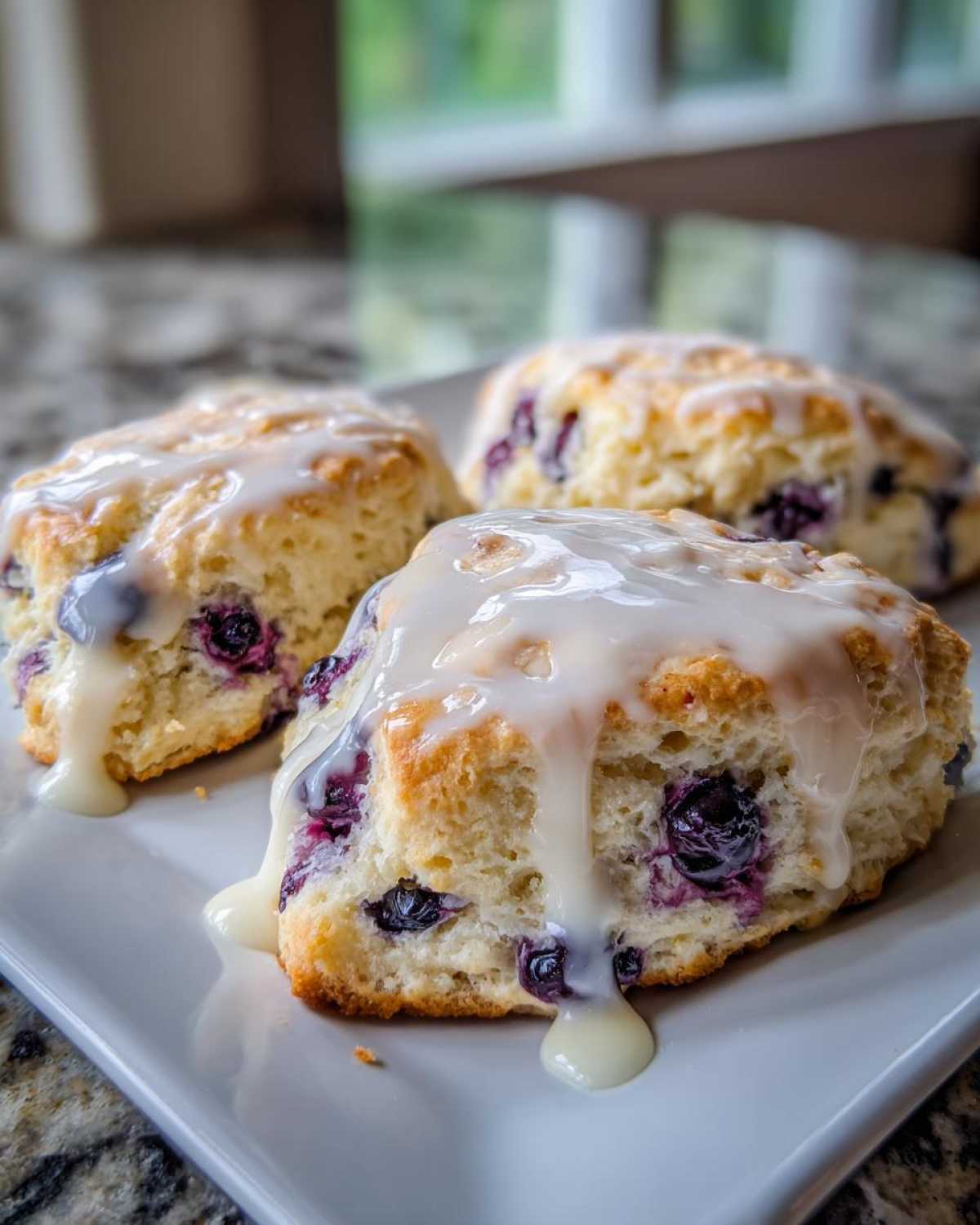 Three freshly baked Blueberry Lemon Scones Recipe topped with thick, dripping lemon glaze on a white plate.