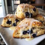 A stack of four golden Blueberry Lemon Scones Recipe triangles drizzled heavily with white lemon glaze.