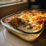 A portion scooped out of a golden-topped chicken pot pie casserole showing creamy filling, chicken, peas, and carrots.