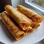 A stack of golden brown, rolled Chicken Taquitos resting on a white plate near a window.
