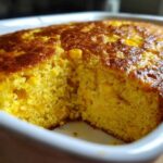 A close-up of a freshly baked, golden brown jiffy corn casserole with visible corn kernels inside a white baking dish.