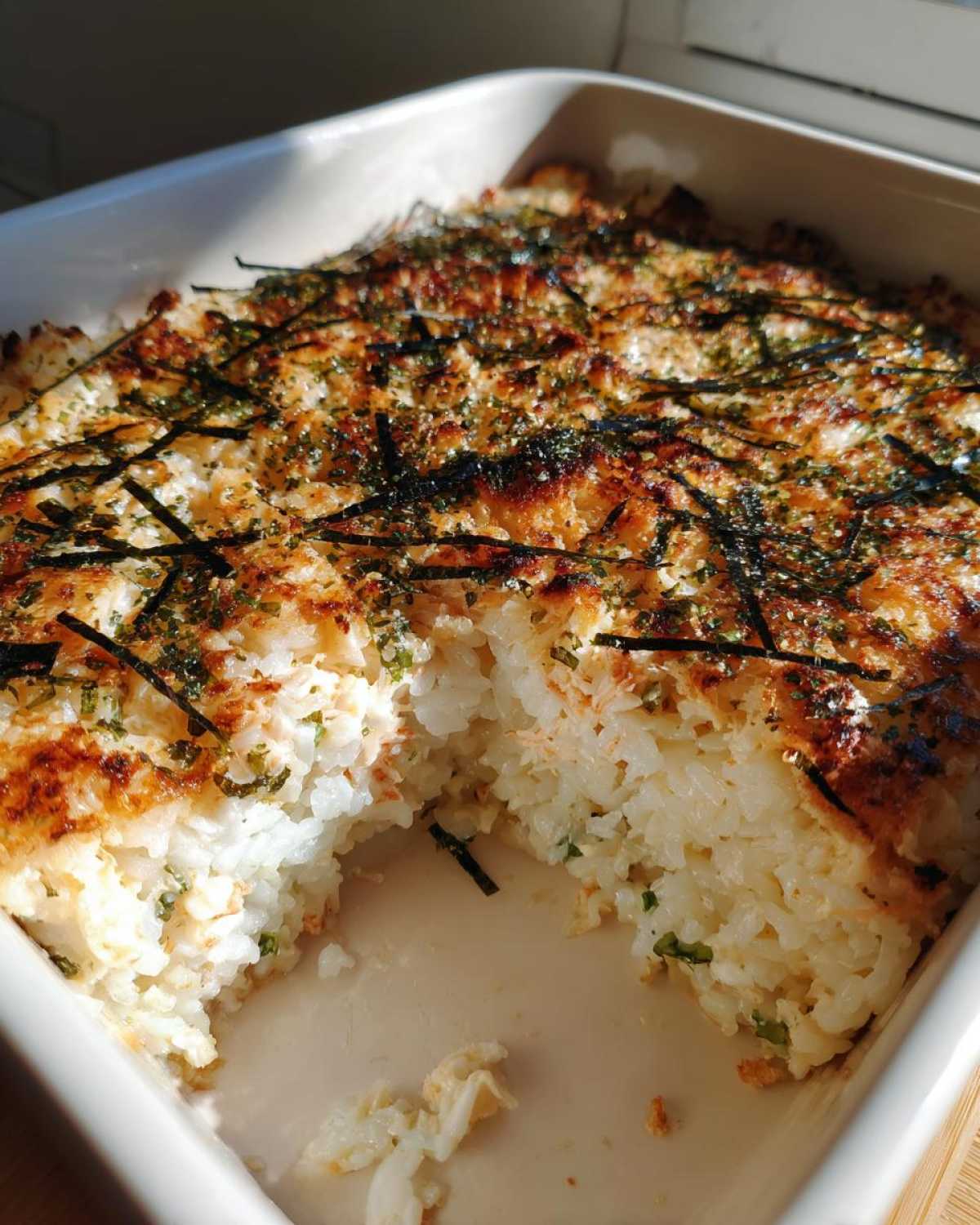 A freshly baked sushi bake casserole in a white dish with a scoop removed, showing the creamy rice texture and crispy nori topping.