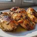 Four pieces of perfectly grilled chicken breasts coated in herbs from the Greek Chicken Marinade, resting on a white plate.