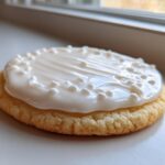 Close-up of a round sugar cookie topped with thick, glossy white sugar cookie icing and white nonpareils.
