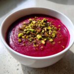 Close-up of a bowl of vibrant, magenta Harissa Beet Soup Recipe topped generously with chopped green pistachios.