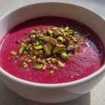 A close-up of a vibrant magenta Harissa Beet Soup Recipe garnished generously with chopped green pistachios.