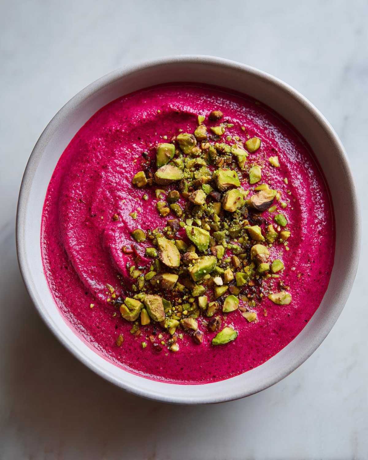 Overhead view of a vibrant magenta Harissa Beet Soup Recipe garnished generously with chopped green pistachios in a light bowl.