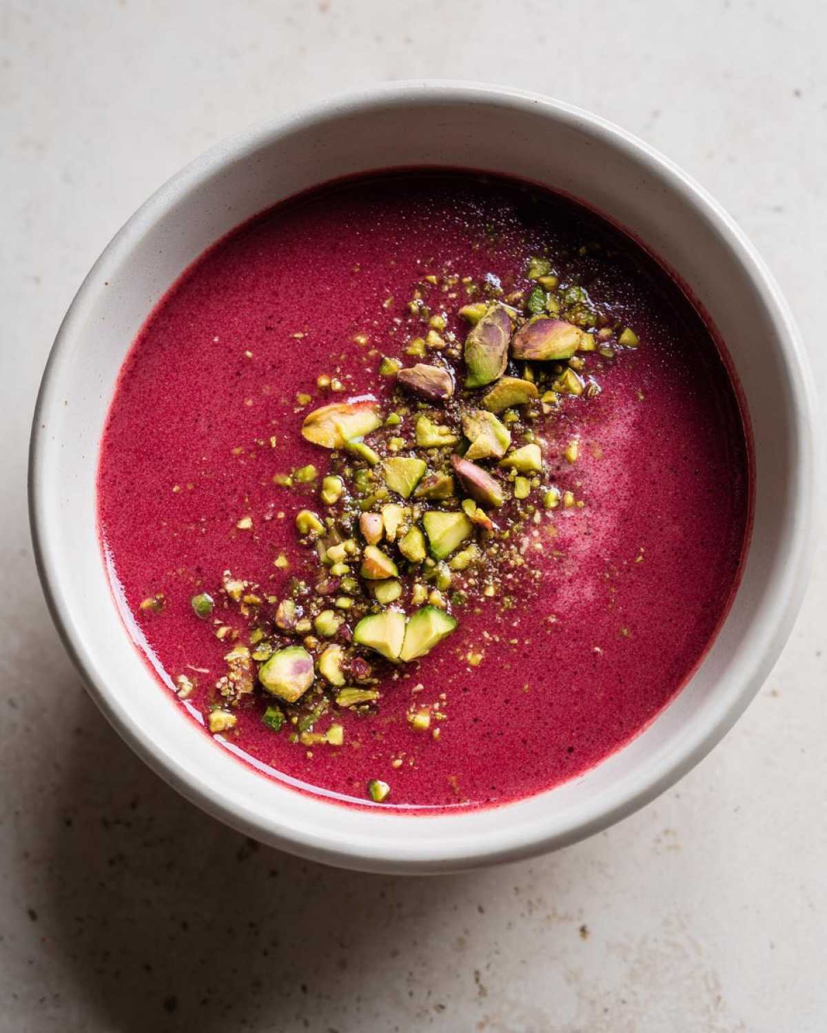 Overhead view of a bowl of vibrant magenta Harissa Beet Soup Recipe garnished generously with chopped green pistachios.