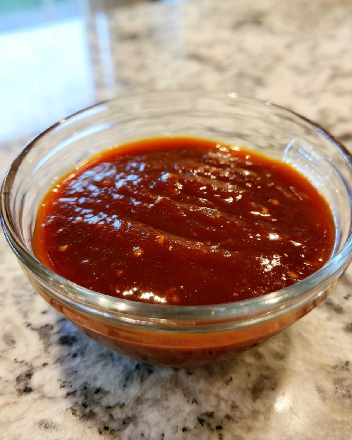 A close-up shot of thick, rich, reddish-brown homemade bbq sauce in a small clear glass bowl.