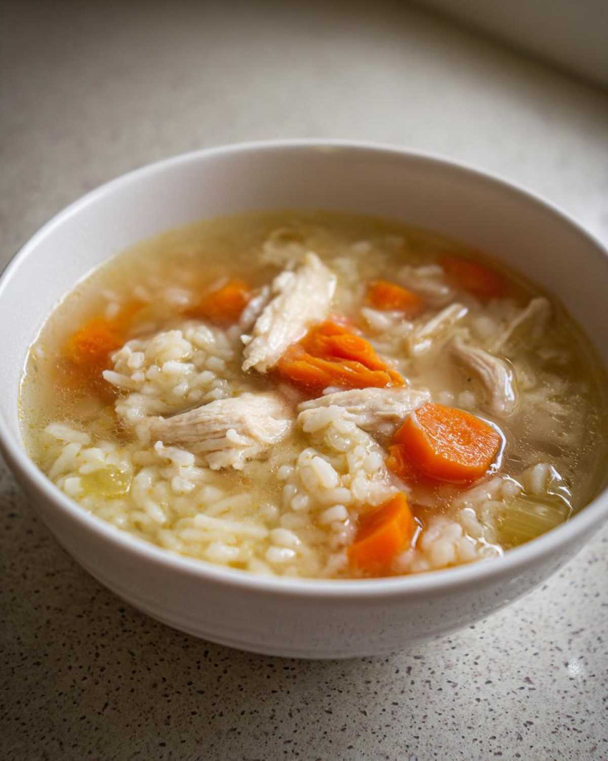 Close-up of a white bowl filled with homemade chicken and rice soup, featuring shredded chicken and bright orange carrots.