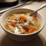 Close-up of a white bowl filled with homemade chicken and rice soup, featuring shredded chicken and bright orange carrots.