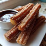 A stack of freshly fried Homemade Churros with Chocolate Sauce, coated in cinnamon sugar, served on a white plate.