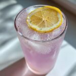 Close-up of a tall glass filled with pink Homemade Lavender Lemonade, ice, and topped with a lemon slice.