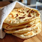 A stack of freshly cooked Homemade Pita Bread from Scratch, wrapped partially in a white cloth on a wooden board.