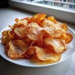 A white plate piled high with golden brown, homemade potato chips dusted lightly with salt.