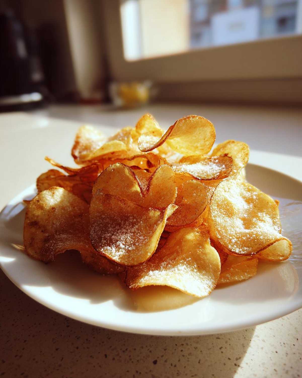 A pile of golden, homemade potato chips lightly dusted with salt, served on a white plate in bright sunlight.