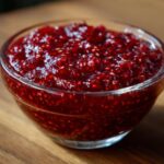 A close-up shot of thick, vibrant red homemade raspberry jam, full of seeds, in a clear glass bowl.