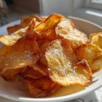 A close-up of freshly made, golden brown potato chips sprinkled with coarse salt, served in a white bowl.