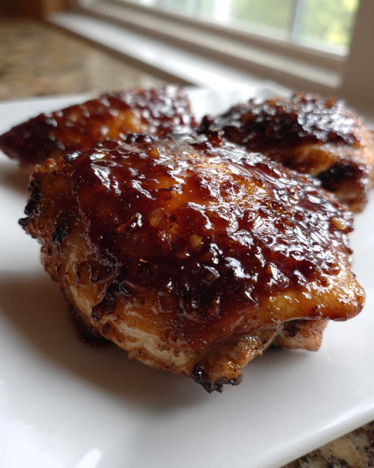 Close-up of three glistening Honey Garlic Chicken Thighs coated in a thick, dark glaze, served on a white plate.