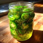 Close-up of vibrant green, sliced candied jalapenos soaking in a sugary syrup inside a clear glass jar.